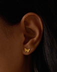 Close-up of a person’s ear wearing Freedom to Soar Butterfly Stud Earrings in 18k gold plate. The dark background highlights the jewelry’s transformative design alongside the person’s dark hair and skin.