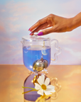 A hand with purple nails lifts the lid of a glass teapot filled with blue tea from the Blue Lotus Tea Ceremony with Zhena. A moon-shaped infuser hangs inside, while a white flower rests on the golden surface against a pastel background.