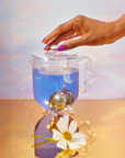 A hand with purple nails lifts the lid of a glass teapot filled with blue tea from the Blue Lotus Tea Ceremony with Zhena. A moon-shaped infuser hangs inside, while a white flower rests on the golden surface against a pastel background.