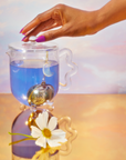 A hand lifts the lid of a glass teapot filled with 999: Divine Completion tea. A moon-shaped infuser hangs inside, while a white flower rests on a reflective gold surface against a pastel sky backdrop.