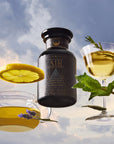 A dark glass jar of Air tea is surrounded by a glass teacup filled with yellow tea and lavender, and a coupe glass with golden tea, mint leaves, and a lemon slice, all set against a cloudy sky.
