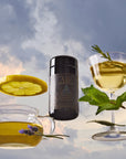 Air, a black cylindrical tea container, is placed between a glass teacup with green tea, lemon, and lavender, and a cocktail glass with tea, mint, rosemary, and lemon slice, all set against a cloudy sky background.