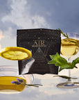 A floating tea package labeled "Air" is surrounded by a teacup, a coupe glass with light tea, a lemon slice, fresh mint, and lavender, all set against a cloudy sky background.