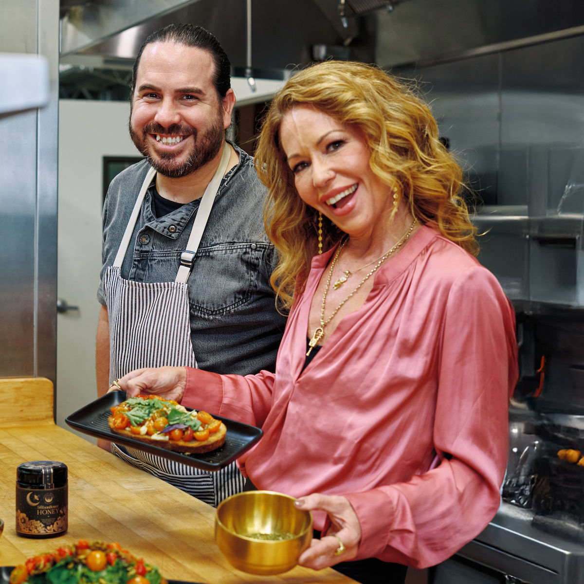 A smiling man in a striped apron and a woman in a pink blouse hold plates of food in a commercial kitchen, preparing and presenting dishes enhanced with Matcha Herbed Salt.