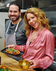 A smiling man in a striped apron and a woman in a pink blouse hold plates of food in a commercial kitchen, preparing and presenting dishes enhanced with Matcha Herbed Salt.