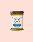 A 3 oz (85 g) jar of Savannah Bee Co. Honey - Acacia, featuring a brown lid, sits against a pastel pink background. The label displays floral illustrations and states it's pure, natural, single-varietal honey from Hungary.