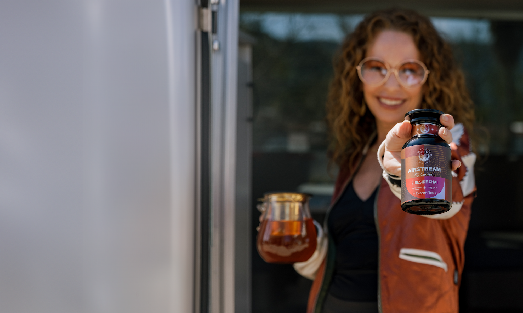 A woman with curly hair and large glasses stands inside an Airstream Trailer, extending a jar of loose leaf tea towards the camera in one hand and holding a pitcher of tea in the other. She is smiling, wearing a brown jacket, and a blurred background is visible through the window.