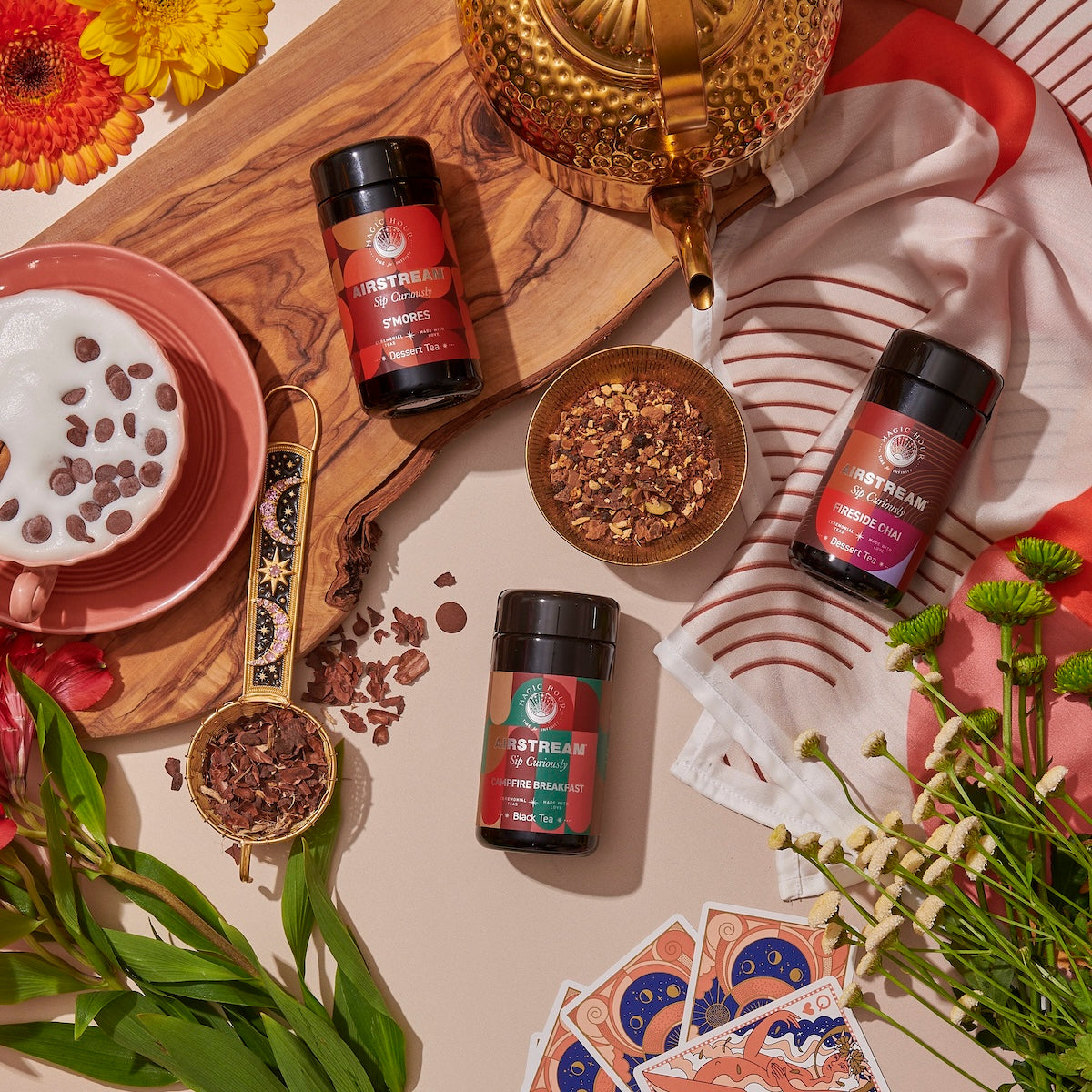 Top view of a cozy tea setup featuring the Sip Curiously Gift Set from Magic Hour, including a wooden board, a gold teapot, three tins of loose tea, an exclusive blends latte in a red cup, the Midas Touch Tea strainer, assorted flowers, playing cards, and a red-and-white patterned cloth. Scattered tea leaves add texture to the scene.