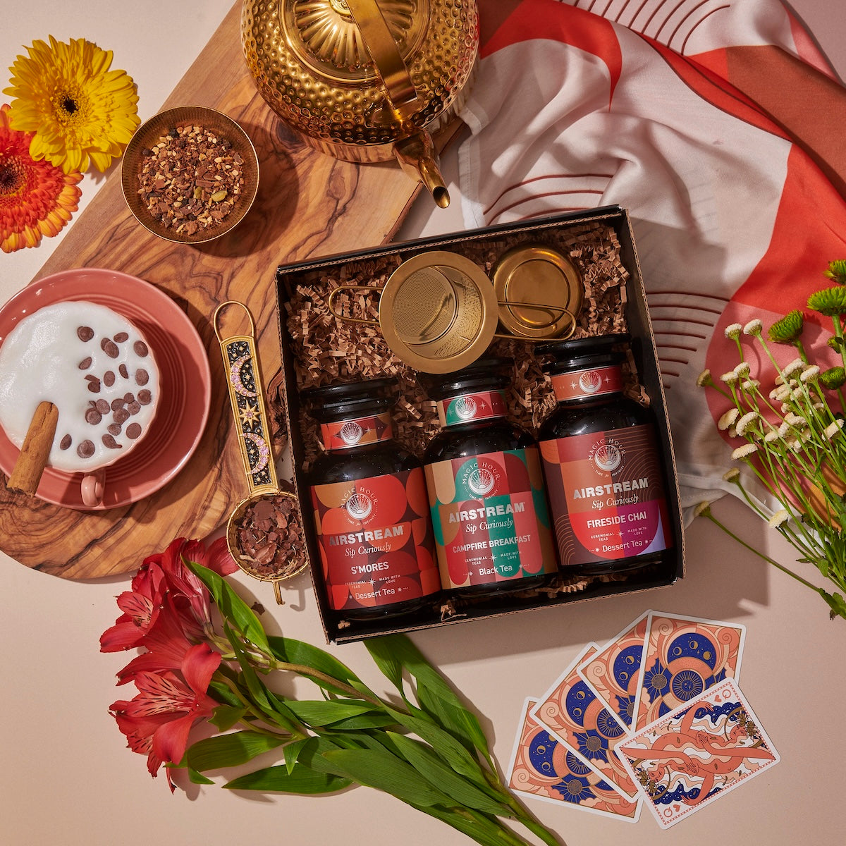 A cozy scene features the Magic Hour's Sip Curiously Gift Set with three tins labeled S'mores, Ginger Breadhouse, and Fireside Chai. Surrounding the box are a bowl of frothy milk with chocolate chips, flowers, a teapot, cards, chocolate shavings, and a colorful cloth.
