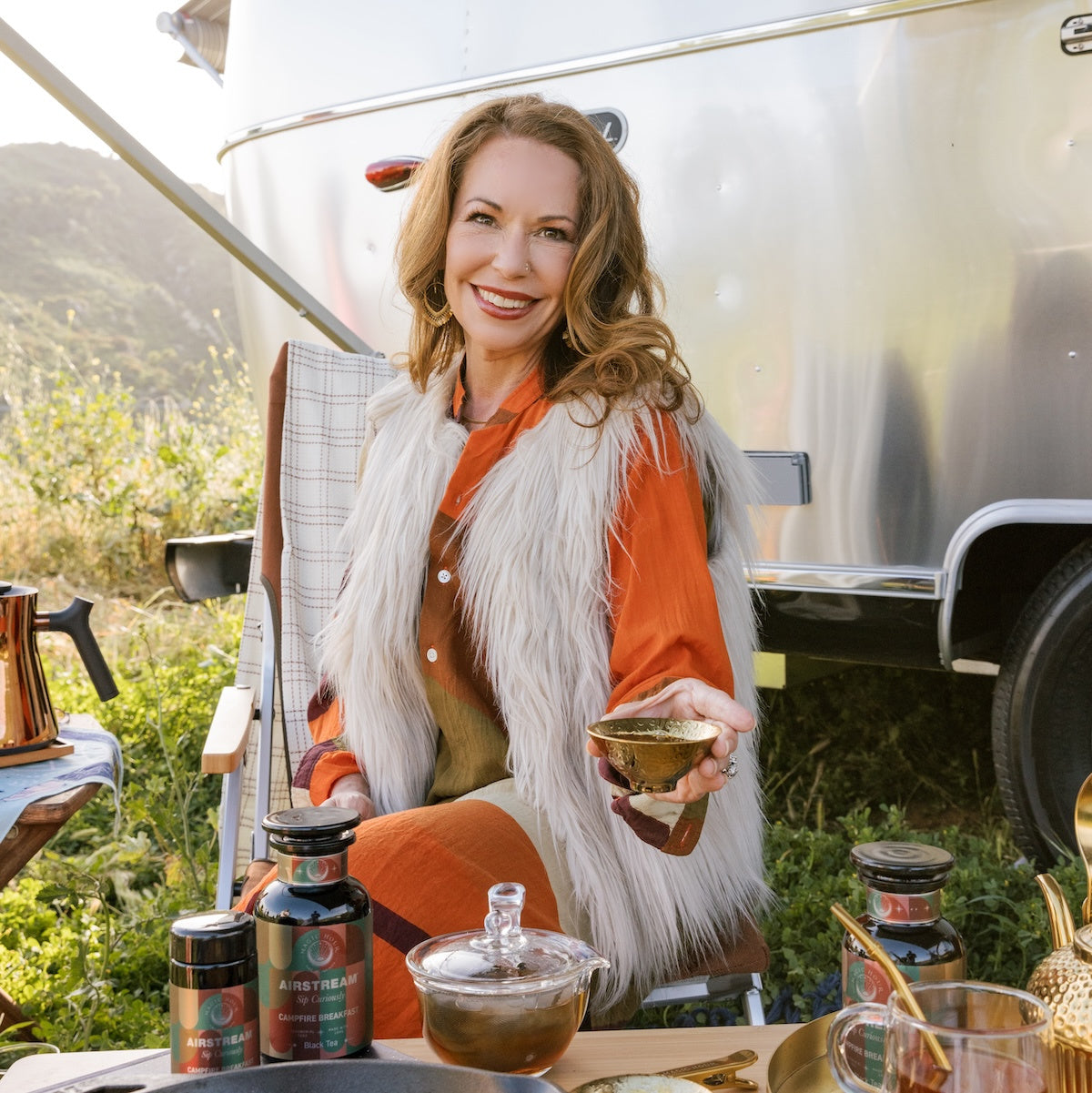 Zhena in a fur vest and orange dress seated outside an Airstream trailer holding a teacup. Various jars and a teapot are on a table in front of her.