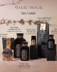 A selection of Magic Hour tea products is arranged artistically against a soft background. Items include various jars, pouches, and packaging in dark, elegant designs. Labels indicate serving capacities and names such as Sampler Pouch and Luxe Refill Pouch, complemented by the luxurious Goddess of Earl Set and Gold Luxe Loose Leaf Tea Strainer.