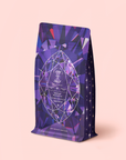 A vibrant purple refill pouch adorned with geometric, gem-inspired designs and star motifs, labeled “Amethyst: Strawberry-Passionfruit Wellness Tea,” stands upright on a pale pink background. Inspired by the calming essence of amethyst and spiritual wellness, this strawberry-passionfruit blend brings a soothing touch of tranquility to your daily ritual.