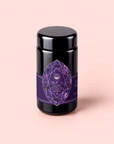 A black jar adorned with a purple geometric crystal label reading “Amethyst: Strawberry-Passionfruit Gemstone Wellness Tea” rests on a pale pink background, evoking spiritual wellness and the calming aura of gemstone-infused tea.