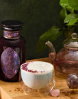 A cozy scene showcases a wooden table with Amethyst: Strawberry-Passionfruit Gemstone Wellness Tea in a glass teapot, paired with a frothy rose petal-topped drink. Complemented by an amethyst crystal and dark jar, a beaded tassel necklace accentuates the pink gem, while greenery enhances the spiritual ambiance.