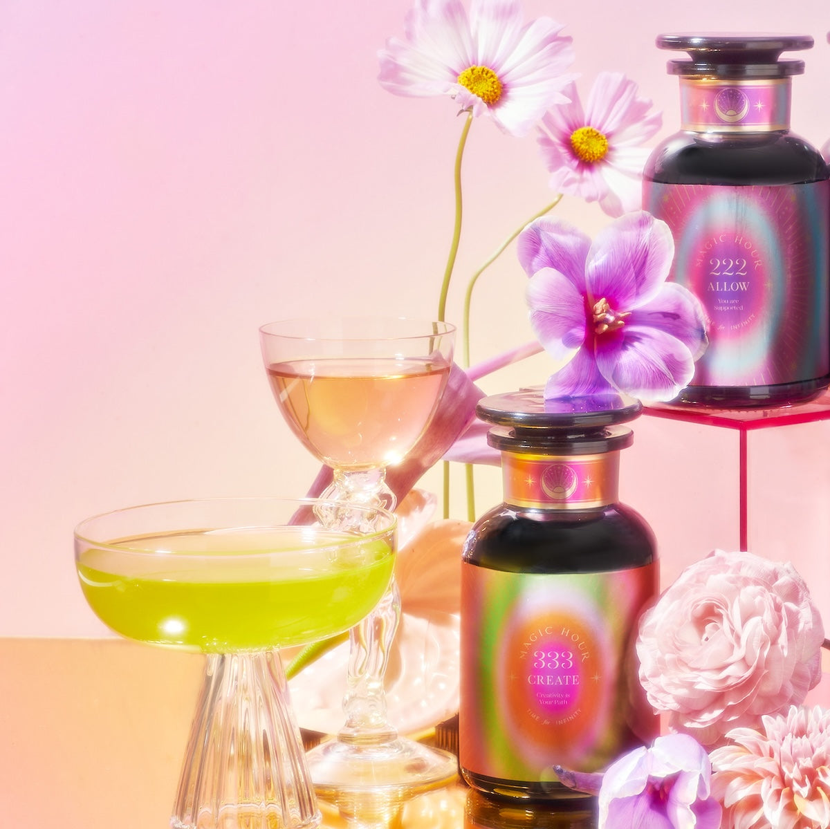 Vibrant potion bottles featuring the Angel Number Tea Trinity with floral labels rest on a reflective surface amid delicate pink and purple flowers, alongside organic loose-leaf teas. Nearby, two elegant cocktail glasses brim with colorful liquids, crafting an enchanting scene.
