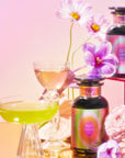 Vibrant potion bottles featuring the Angel Number Tea Trinity with floral labels rest on a reflective surface amid delicate pink and purple flowers, alongside organic loose-leaf teas. Nearby, two elegant cocktail glasses brim with colorful liquids, crafting an enchanting scene.