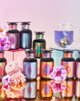 Eight iridescent perfume bottles, pastel flowers, a glass teacup of blue Angel Tea, and a gold tea infuser are displayed on mirrored platforms with a pastel gradient backdrop for the Angel Tea Journey Subscription.