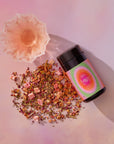 A pink elixir in a delicate glass sits beside a black jar labeled "333: Mango-Lemon Balm-Rose Herbal Tea with Passionflower," with loose tea leaves and dried fruit scattered nearby, inspiring creativity and heart-centered expression.