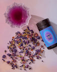 A glass of purple herbal tea, a dark container labeled "444: Sachiel's Prosperity," and scattered apple-berry, butterfly pea flower, and blue spirulina blend evoke the mystique of a blue elixir on a light pink surface.