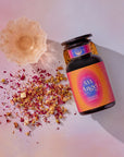 A glass jar labeled "555: Celestial Harmony: Papaya-Mango-Rose Tea with Ashwagandha" rests on a pink surface beside a dish and vibrant dried petals, evoking stress relief and the soothing magic of this unique tea blend in a dreamy, magical scene.