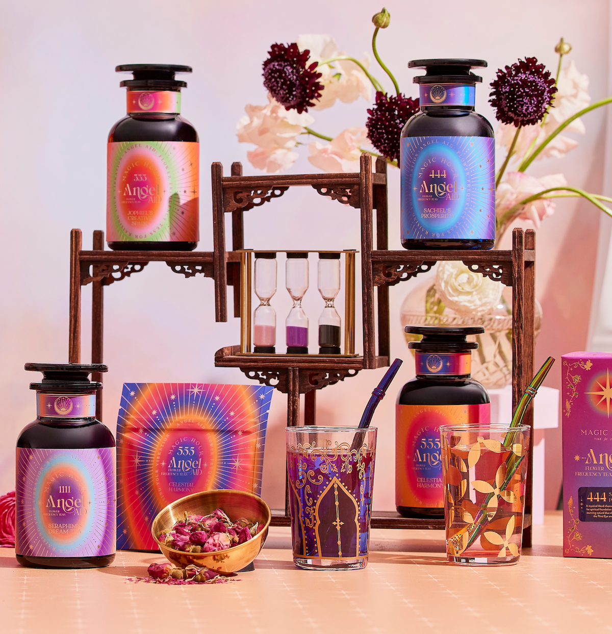 A vibrant display of Angel Tea Journey Subscription bottles, glasses, a gold bowl with rose petals, hourglasses, and decorative boxes on a wooden stand with flowers in the background evokes a spiritual tea ritual experience.