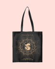 A black cotton tote bag named the Magic Hour Tote Bag showcases "Magic Hour" at the top, with a decorative number 5 surrounded by a floral and sunburst design. Below this, a metallic gold logo states "Celebrating 5 Years of Magic," all set against a soft pink background.
