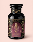 A dark glass jar of Magic Hour tea labeled “Aphrodite’s Alchemy: Damiana & Rose White Tea,” featuring an illustration of the Greek goddess of love seated on a lotus, with rose motifs and gold decorative details set against a soft pink background.