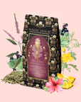 The "Aphrodite's Alchemy - Damiana & Rose White Tea" package showcases an illustration of a woman inspired by Aphrodite, the Greek goddess of love, encircled by herbs, hibiscus, pineapple, blueberries, and yellow flowers set against a pink background.