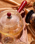 The Swivel Side Pour Teapot for Tea Ceremony, with its elegant borosilicate glass body, wooden handle and lid, sits gracefully on a tray among glass cups, a jar labeled “Magic Honey,” dried herbs, and cinnamon sticks atop golden fabric. Reminiscent of a Japanese kyusu teapot, this piece seamlessly blends tradition with modern simplicity to create an inviting tea experience.