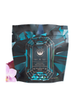 A colorful packaging of "Magic Hour Rest, Relaxation & Sleep Sampler Set" featuring a large gemstone graphic. The package is designed in hues of blue, green, and teal. An orchid flower is partially visible next to the vibrant loose leaf tea package.