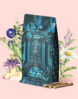 A jewel-toned tea package labeled "Aquamarine Dream - Soothing Herbal Ayurvedic Adrenal Tonic Refill Pouch" stands in front of illustrated flowers, apple slices, and licorice roots, all set against a pale pink background. The gem-like blue design is inspired by the calming aquamarine tea inside—a soothing herbal blend crafted to ease the senses.