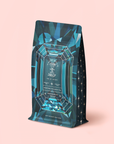 A blue geometric tea bag package labeled "Aquamarine Dream - Soothing Herbal Ayurvedic Adrenal Tonic Refill Pouch" sits upright on a pale pink background, adorned with star patterns and gemstone-inspired designs. This aquamarine blend is a calming herbal infusion designed to soothe the senses and promote peaceful evenings.