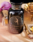 A black glass jar labeled "Aquarius: Visionary Goddess Tea" with gold and floral designs sits on gold satin, surrounded by a purple calla lily, dried herbs, gold-wrapped chocolates, and Organic Golden Monkey Tip Black Tea leaves.