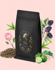 A black bag of tea labeled "Aries - Garden of Eden Oolong" sits among loose tea leaves, a pink rose, blackberries on a branch, and two green bergamot fruits against a light pink background—a dreamy scene for this oolong blend.