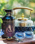 A jar of "Artemis of the Wilds: Wild Moon White Tea," a glass teapot with golden accents, and two glass teacups filled with blue tea are displayed outdoors amid lush greenery and a delicate blue flower. This enchanting low-caffeine blend transforms in hue with a splash of citrus, creating a magical tea experience.