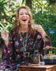 A woman in a floral dress sits outdoors, smiling as she holds a small cup of Artemis of the Wilds: Wild Moon White Tea. She’s surrounded by greenery, with a teapot, bottle, and glass bowl arranged on the wooden table in front of her, savoring the delicate flavor of this low-caffeine blend.