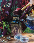 A hand pours Artemis of the Wilds: Wild Moon White Tea from a blue glass teapot into two small cups on a wooden table, with the floral tea jar and fresh herbs nearby. The person is dressed in a floral-patterned garment.