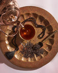A copper teapot is pouring Magic Hour Assam Black Tea into a glass teacup placed on an ornate, round, golden tray. Next to the teacup, there is a pile of loose tea leaves known for their malty flavor and full-bodied character, a metal scoop, and a string of black beads with a tassel. The background is white with a soft shadow.