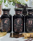 Three black jars labeled "Aries," "Taurus," and "Gemini" sit on a white surface with a gold tin, ornate red candle, and beads nearby. Each jar, perfect for storing Magic Hour's organic loose leaf tea, displays a constellation-themed design in light pink and gold. Flowers and a cup are visible in the background.