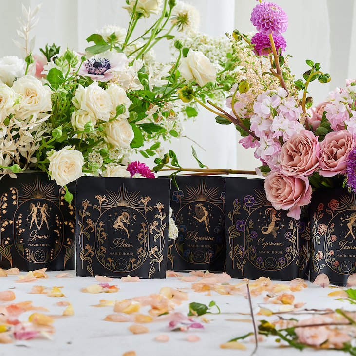 A display of ornamental Astrology Sampler Tea Set by Magic Hour featuring floral designs, each labeled with zodiac signs like Pisces, Gemini, Virgo, and Capricorn. The tea set is surrounded by fresh flowers and scattered petals on a white surface, blending celestial charm with delicate health influences.