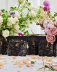 A display of ornamental Astrology Sampler Tea Set by Magic Hour featuring floral designs, each labeled with zodiac signs like Pisces, Gemini, Virgo, and Capricorn. The tea set is surrounded by fresh flowers and scattered petals on a white surface, blending celestial charm with delicate health influences.