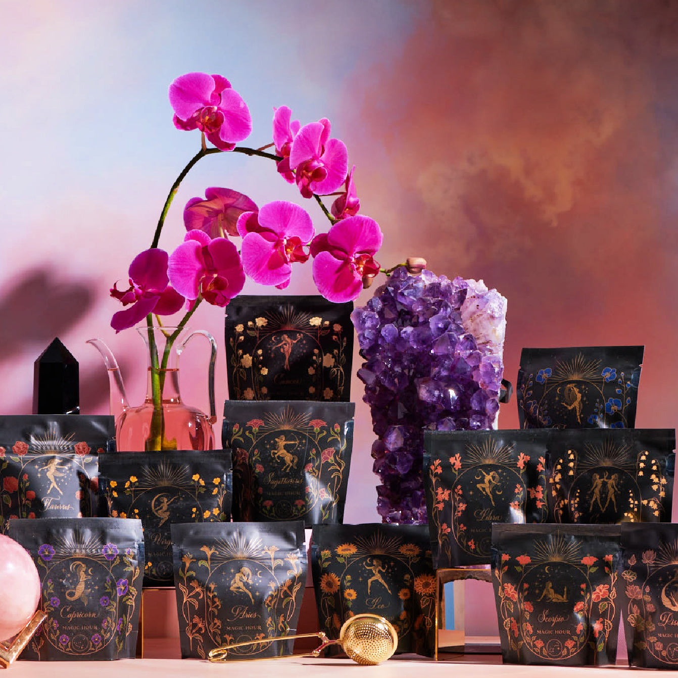 A selection of colorful, intricately designed tea packages from Magic Hour's Astrology Sampler Tea Set are elegantly arranged on a table. In the background, a vibrant pink orchid and a large amethyst crystal cluster stand out against a dreamy, multicolored sky-like backdrop. A golden tea strainer is placed in front.