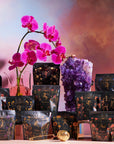 A selection of colorful, intricately designed tea packages from Magic Hour's Astrology Sampler Tea Set are elegantly arranged on a table. In the background, a vibrant pink orchid and a large amethyst crystal cluster stand out against a dreamy, multicolored sky-like backdrop. A golden tea strainer is placed in front.