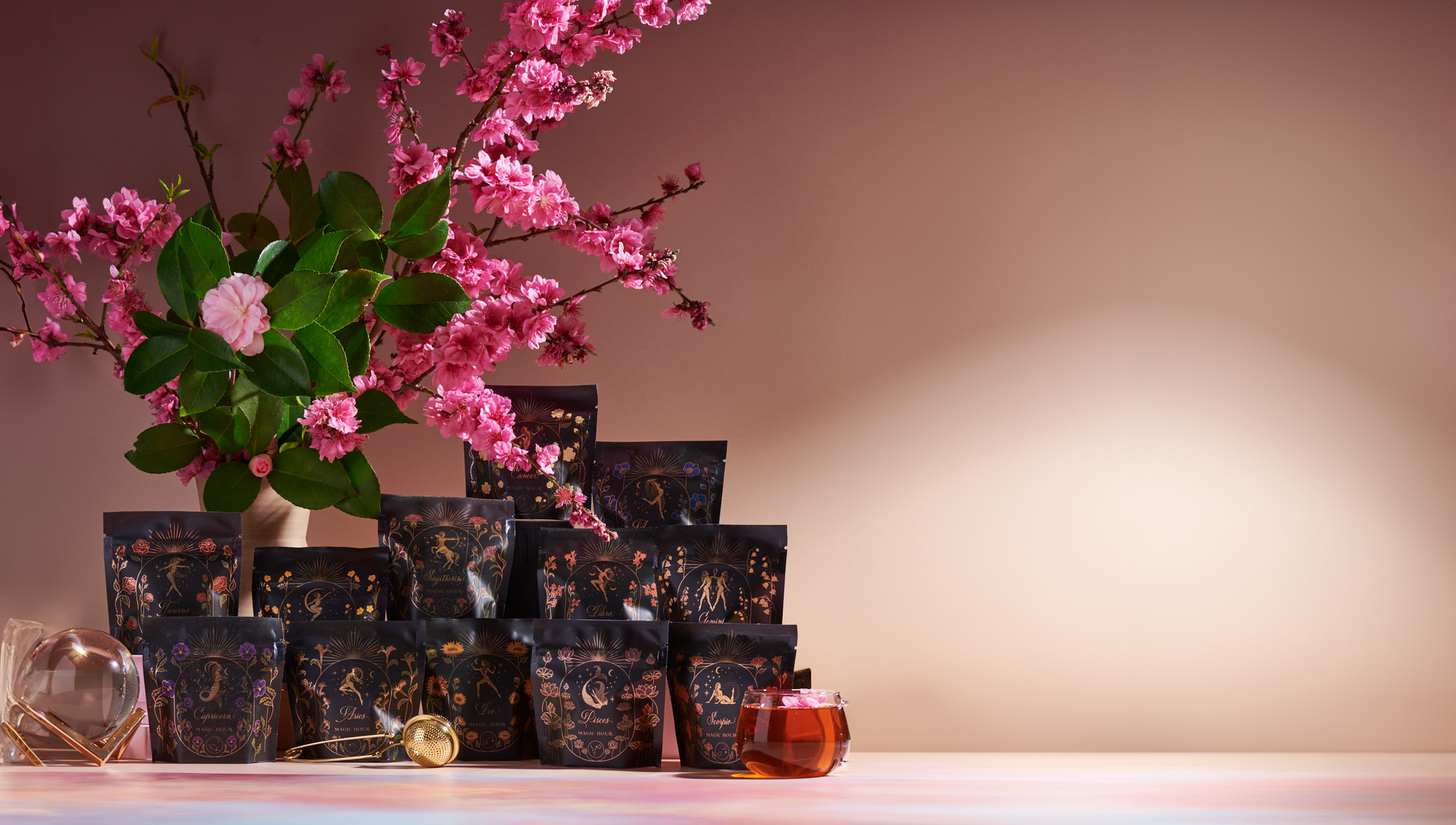 A display of tea packages arranged neatly beside a vase with blooming cherry blossoms. A clear glass mug with tea and a gold tea strainer are placed in the foreground. The background is a soft pastel color.