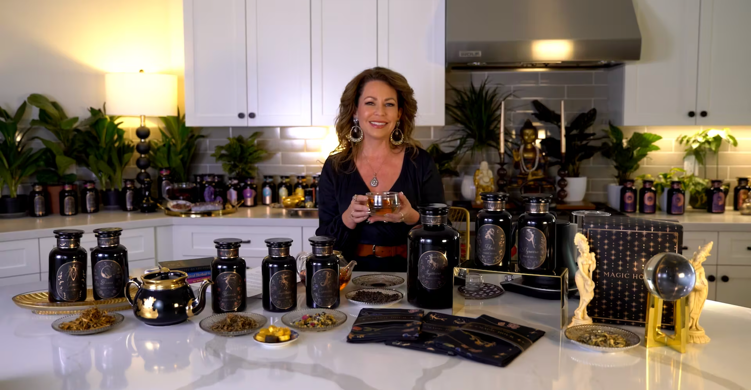 A woman with long hair stands in a modern kitchen filled with various black jars and bottles of herbs and spices. She holds a jar with a smile. The countertop displays additional jars, bowls, and a crystal ball. The background shows more jars and decorative plants.