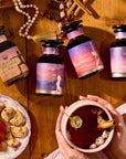 Four Balancing Dosha Bundle wellness tea jars with pastel labels sit on a wooden table. Below, hands hold a cup of spiced, caffeine-free tea beside sliced fruit, biscuits, cinnamon sticks, and beads for a cozy setup.