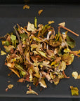 A close-up image of loose-leaf Bai Mudan White Tea by Magic Hour. The handcrafted blend includes various dried leaves and stems in shades of green, brown, and yellow, spread across a dark surface. The white tea leaves appear to be a mix of different sizes and shapes.