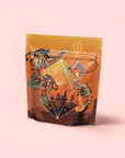 A standing resealable coffee bag with an orange and brown gradient, illustrated monkeys, leaves, and geometric shapes on a pink background. The text reads "Bali Bliss: Coconut-Lime & Ginger-Melon White Tea," inspired by vibrant Bali tea traditions.