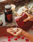 A pair of hands pours Pink Tourmaline Bath Soak from a red, heart-themed pack into one palm over a bathtub adorned with rose petals. Nearby, a black jar of organic tea seed oil and a decorative teapot with cups are placed on the edge of the tub, alongside a lit candle.