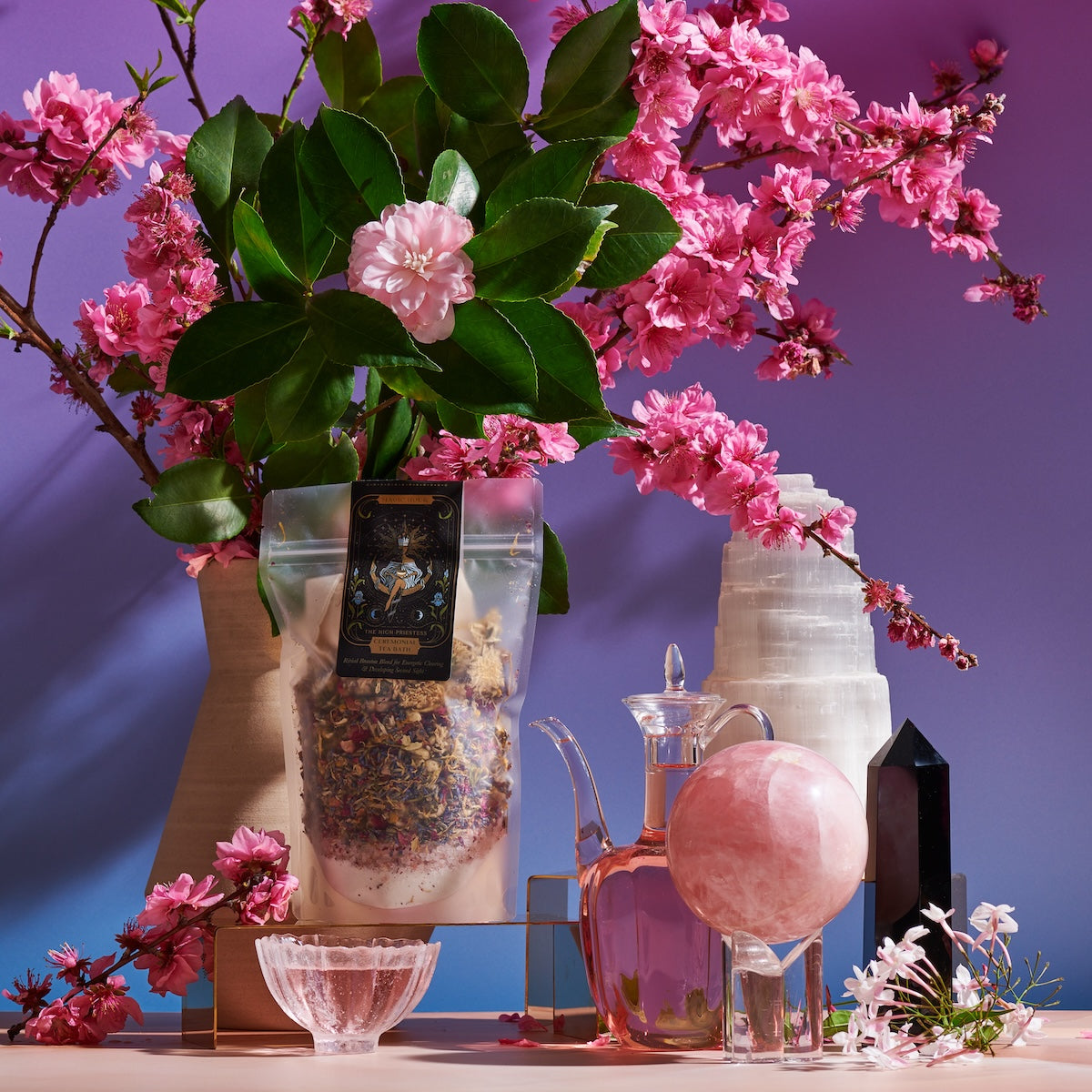 A serene still-life arrangement featuring The High Priestess Box, including a vase with pink blossoms and green leaves, a clear bag of High Priestess Wisdom Tea, a glass teapot, a pink glass orb, various crystals, and a small bowl, all set against a gradient blue-to-purple background.